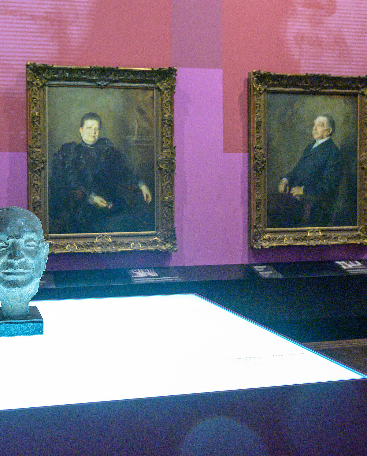 Ausstellungsansicht „Bildgeschichten“, Familie Bernheimer mit Fanny, Lehmann und Otto, im Vordergrund eine Büste, dahinter an der Wand zwei großformatige Porträts, daneben stehend eine Besucherin, Foto: Daniel Schvarcz / Jüdisches Museum München