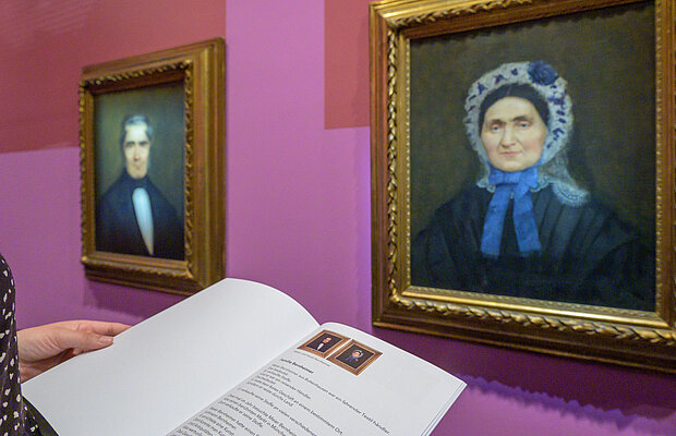 Rundgang durch die Ausstellung „Bild·geschichten“ in leicht verständlicher Sprache, man sieht die aufgeschlagene Begleit·broschüre in Leichter Sprache in der Ausstellung vor zwei Porträts, Foto: Daniel Schvarcz / Jüdisches Museum München 