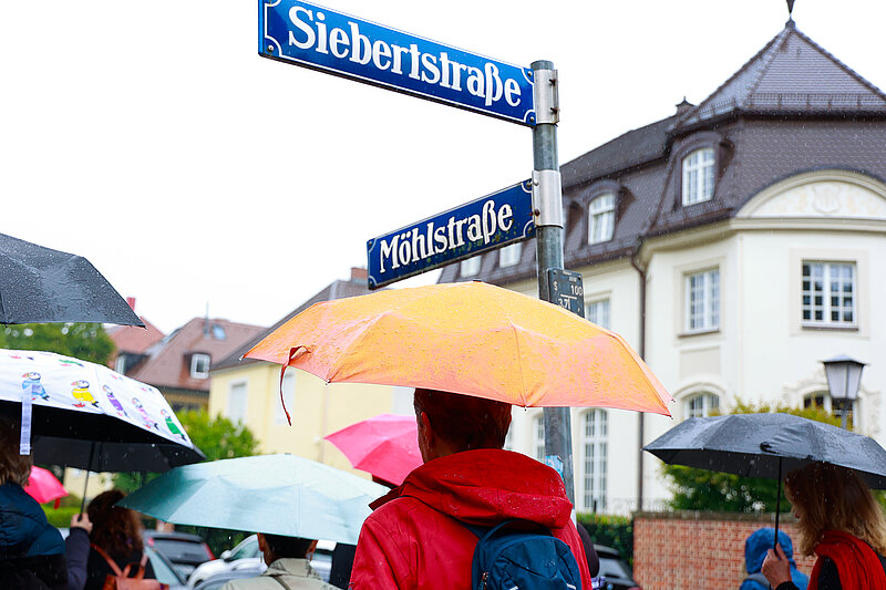 Rundgang durch die Möhlstraße im September, Foto: Marina Maisel Menschen mit bunten Regenschirmen laufen an den Straßenschildern "Möhlstraße" und "Siebertstraße" vorbei