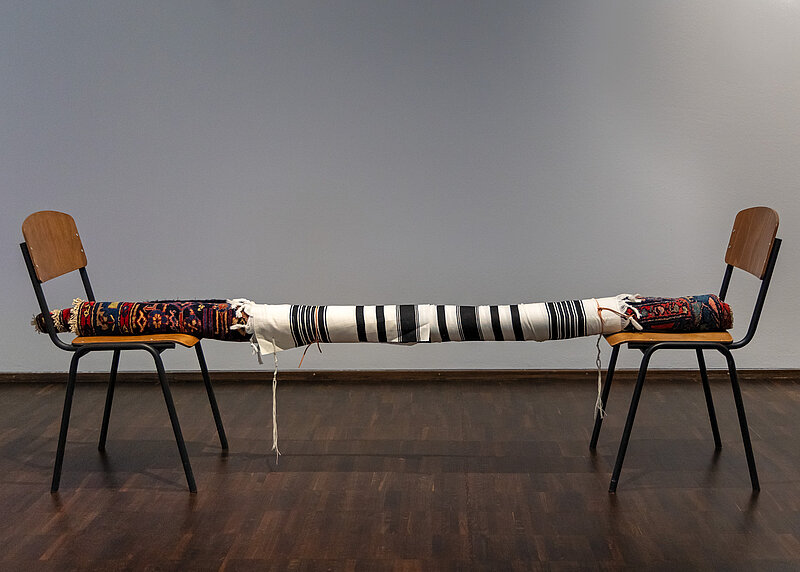 A carpet, a prayer shawl and a leather strap suspended between two chairs 