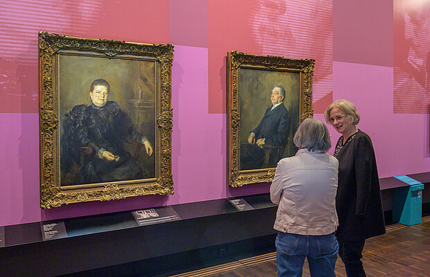 Besucherinnen beim Rundgang durch die Ausstellung „Bildgeschichten“, sie stehen vor zwei Lenbach-Portraits von Fanny und Lehmann Bernheimer, die an einer Wand in Rosetönen hängen, Foto: Daniel Schvarcz / Jüdisches Museum München 