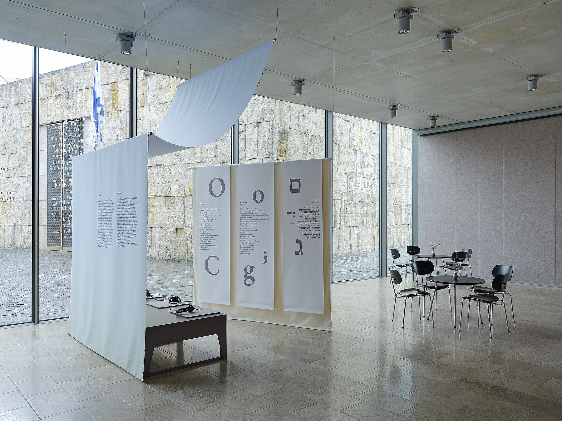 Installation view  "October 7" on display are a small audio installation with seating and text panels hanging from the ceiling on the right in Latin and Hebrew script, photo: Eva Jünger / Jewish Museum Munich 