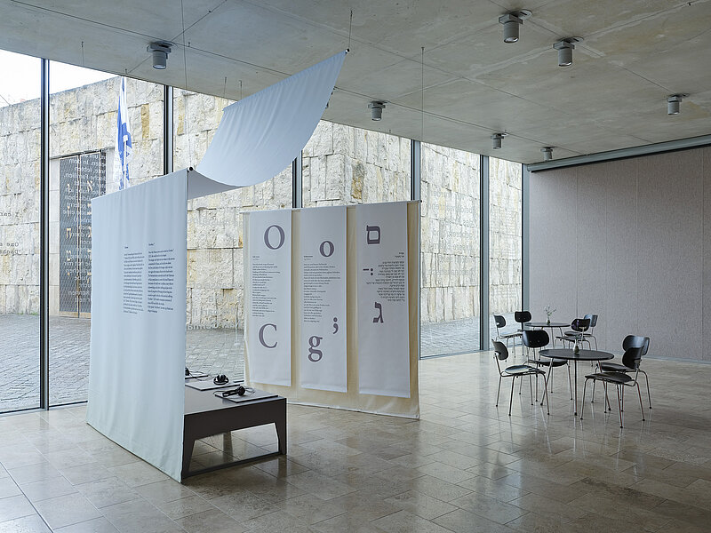 Installation view  "October 7" on display are a small audio installation with seating and text panels hanging from the ceiling on the right in Latin and Hebrew script, photo: Eva Jünger / Jewish Museum Munich 