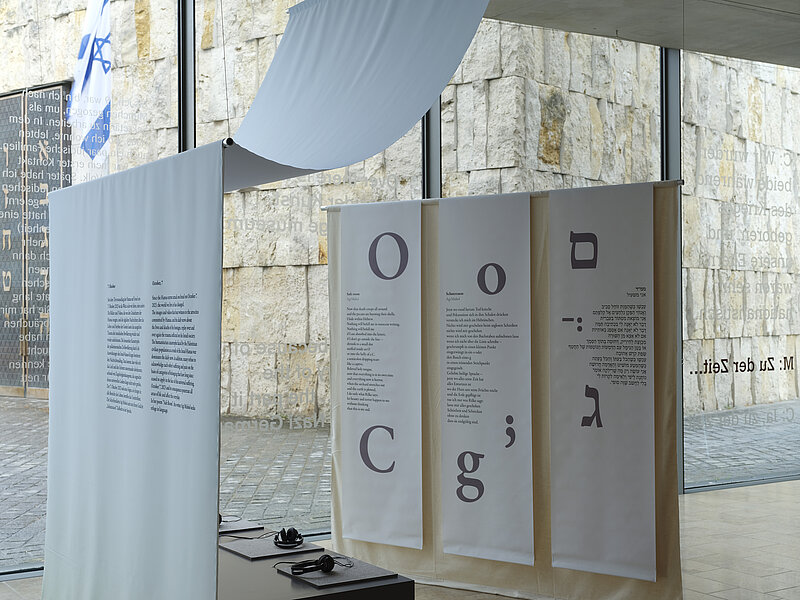 Installation view  "October 7" on display are a small audio installation with seating and text panels hanging from the ceiling on the right in Latin and Hebrew script, photo: Eva Jünger / Jewish Museum Munich 