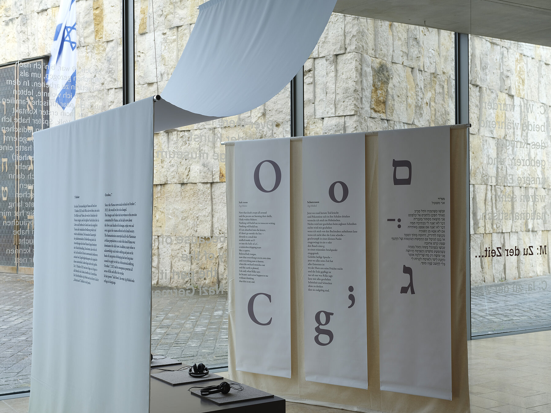 Installation view  "October 7" on display are a small audio installation with seating and text panels hanging from the ceiling on the right in Latin and Hebrew script, photo: Eva Jünger / Jewish Museum Munich 