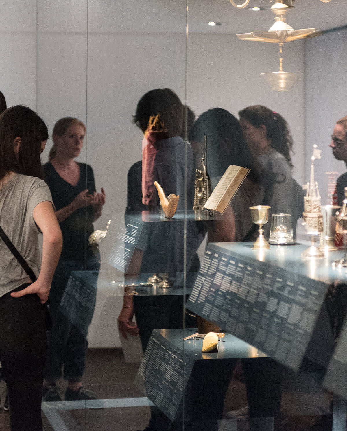 Foto eines Ausstellungsbereichs. Um eine große Vitrine mit metallischen Gegenständen und einem Buch ist eine Gruppe versammelt, die eine Führung erhält