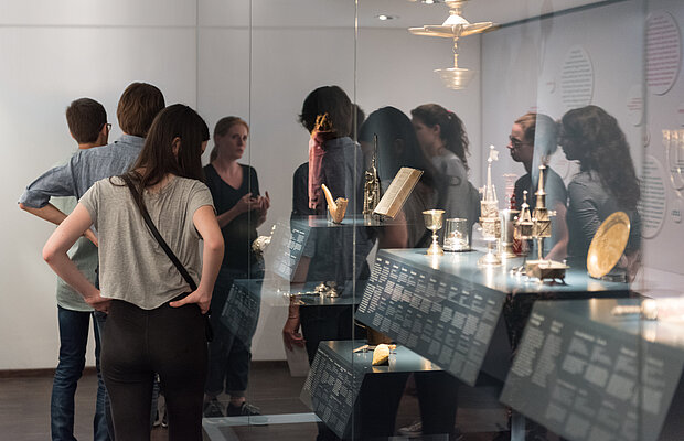 Foto eines Ausstellungsbereichs. Um eine große Vitrine mit metallischen Gegenständen und einem Buch ist eine Gruppe versammelt, die eine Führung erhält