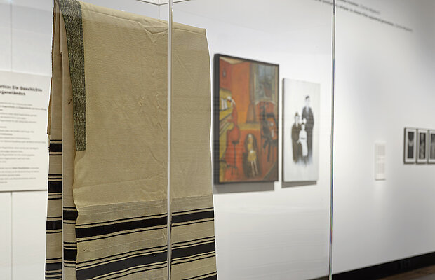 Objekte in der Ausstellung „Die Dritte Generation. Der Holocaust im familiären Gedächtnis“. Im Vordergrund ist ein Talli (Gebetsschal) zu sehen, im Hintergrund zwei Gemälde von Lola Carr. Foto: Eva Jünger / Jüdisches Museum München 