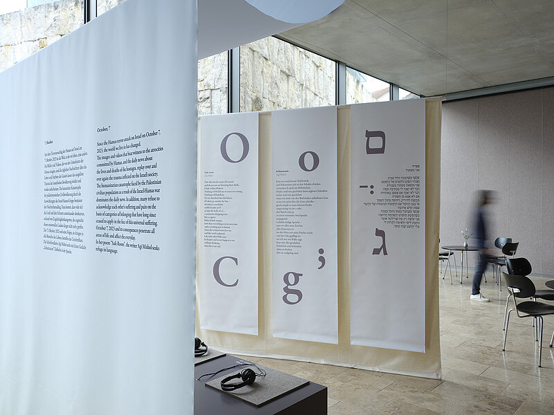 Installation view  "October 7" on display are a small audio installation with seating and text panels hanging from the ceiling on the right in Latin and Hebrew script, photo: Eva Jünger / Jewish Museum Munich 