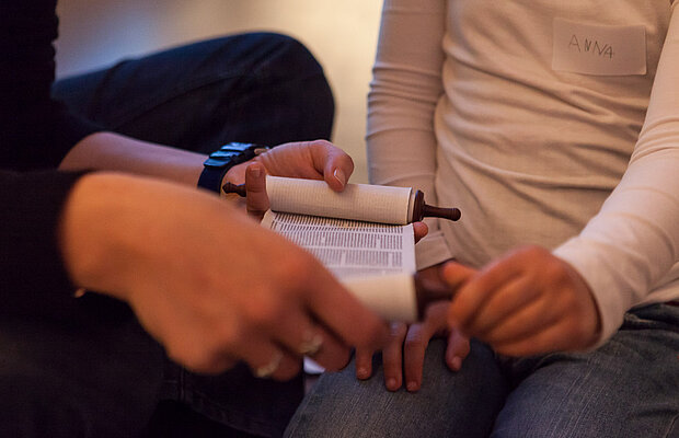 Eine Besucherbetreuerin sitzt in der Dauerausstellung neben einem Kind und zeigt eine kleine Thorarolle. 