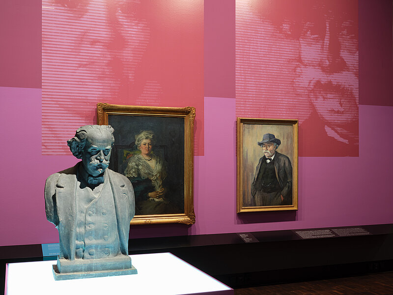 Ausstellungsansicht „Bildgeschichten. Münchner Jüdinnen und Juden im Porträt“ - eine üste im Vordergrund und zwei Porträts im Hintergrund hängen nebeneinander in einem Ausstellungsraum in Rosétönen, große Blowups spiegeln das Motiv der Porträts, Foto: Eva Jünger / Jüdisches Museum München 