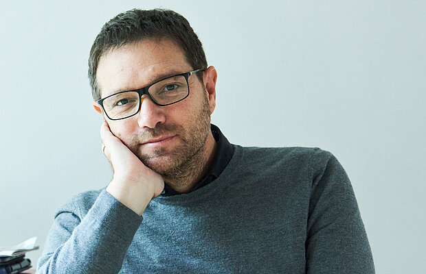 Noam Zadoff sitzt neben einem Schreibtisch und stützt dass Kind in seine Hand.