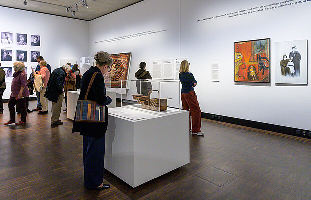 Besucher*innen in der Ausstellung "Die Dritte Generation" betrachten Objekte und Gemälde. 