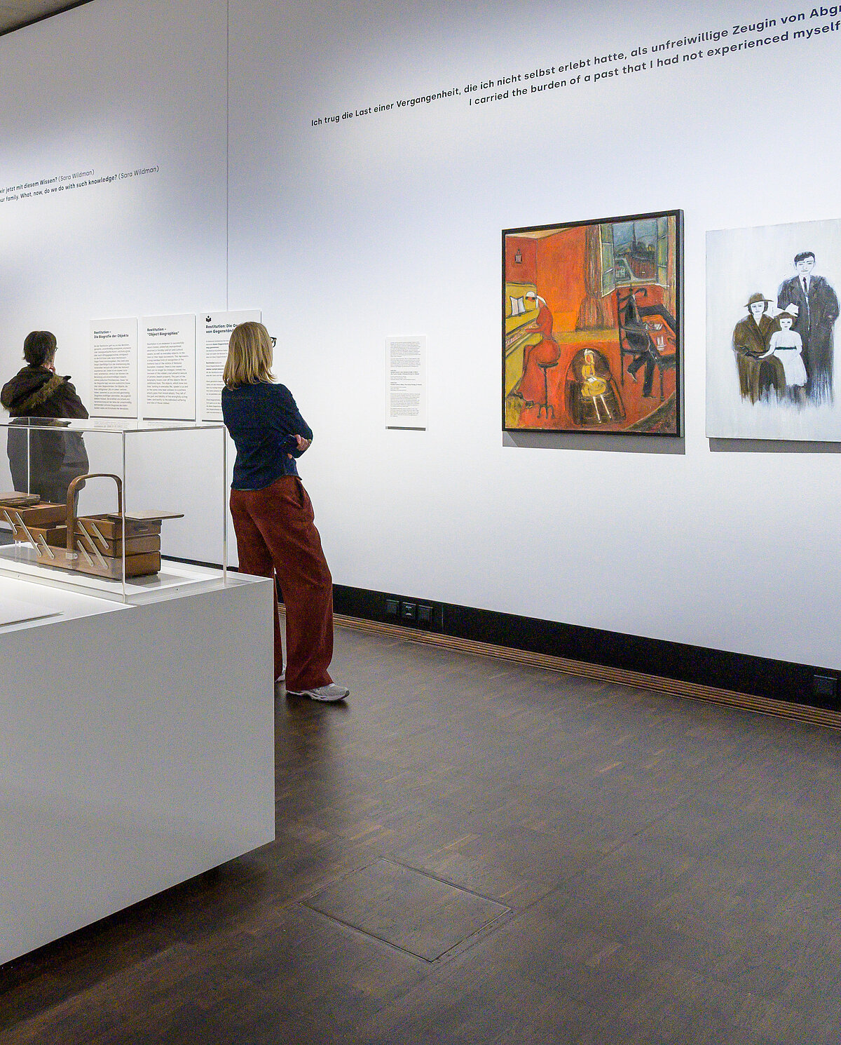 Besucher*innen in der Ausstellung "Die Dritte Generation" betrachten Objekte und Gemälde. 