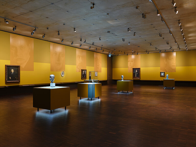 Ausstellungsansicht „Bildgeschichten. Münchner Jüdinnen und Juden im Porträt“ - mehrere Porträts hängen nebeneinander in einem Ausstellungsraum in Gelbtönen, große Blowups spiegeln das Motiv der Bilder, Foto: Eva Jünger / Jüdisches Museum München 