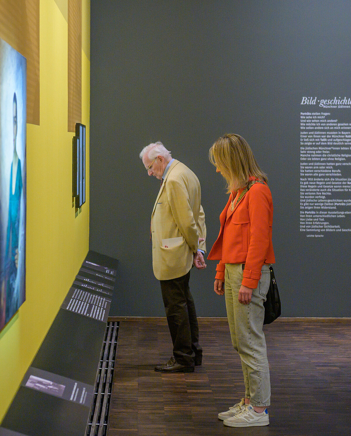Zwei Besucher*innen in der Ausstellung  „Bildgeschichten“ betrachten zwei an einer hellen gelben Wand hängede Gemälde, Foto: Daniel Schvarcz / Jüdisches Museum München 